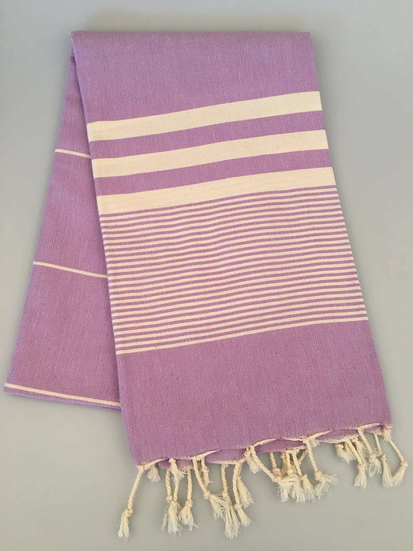 Lavender Turkish towel peshtemal with cream stripes and fringed edges, laid flat on a light background.