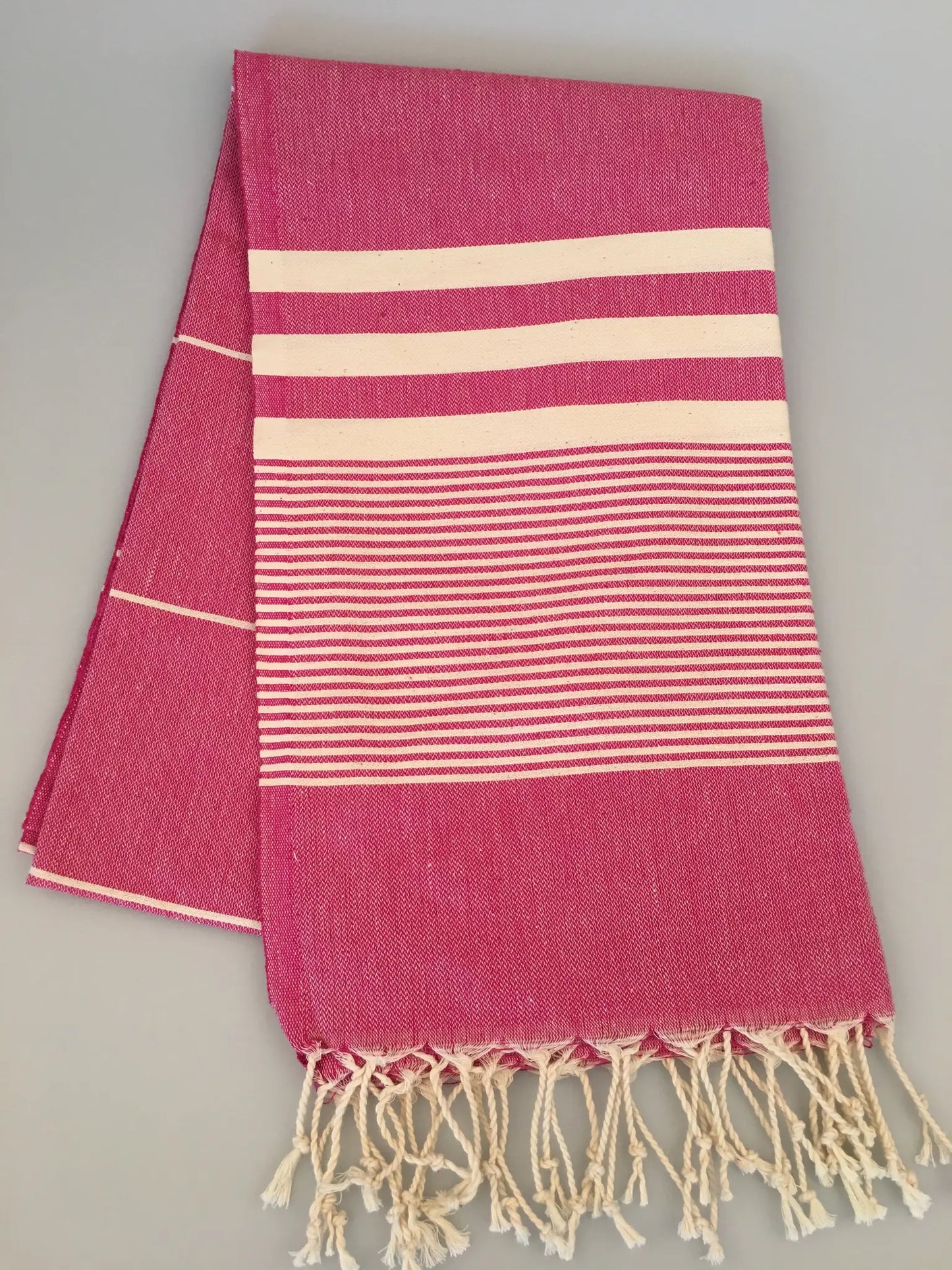 "Pink Turkish towel with white stripes and fringed edges, laid flat on a light gray background."