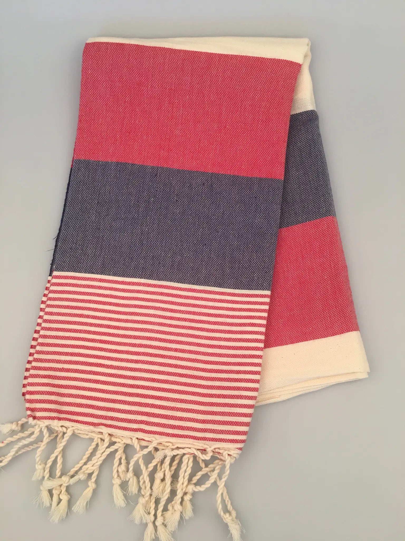 "Red and navy striped Turkish towel with cream fringes, folded on a light gray surface."