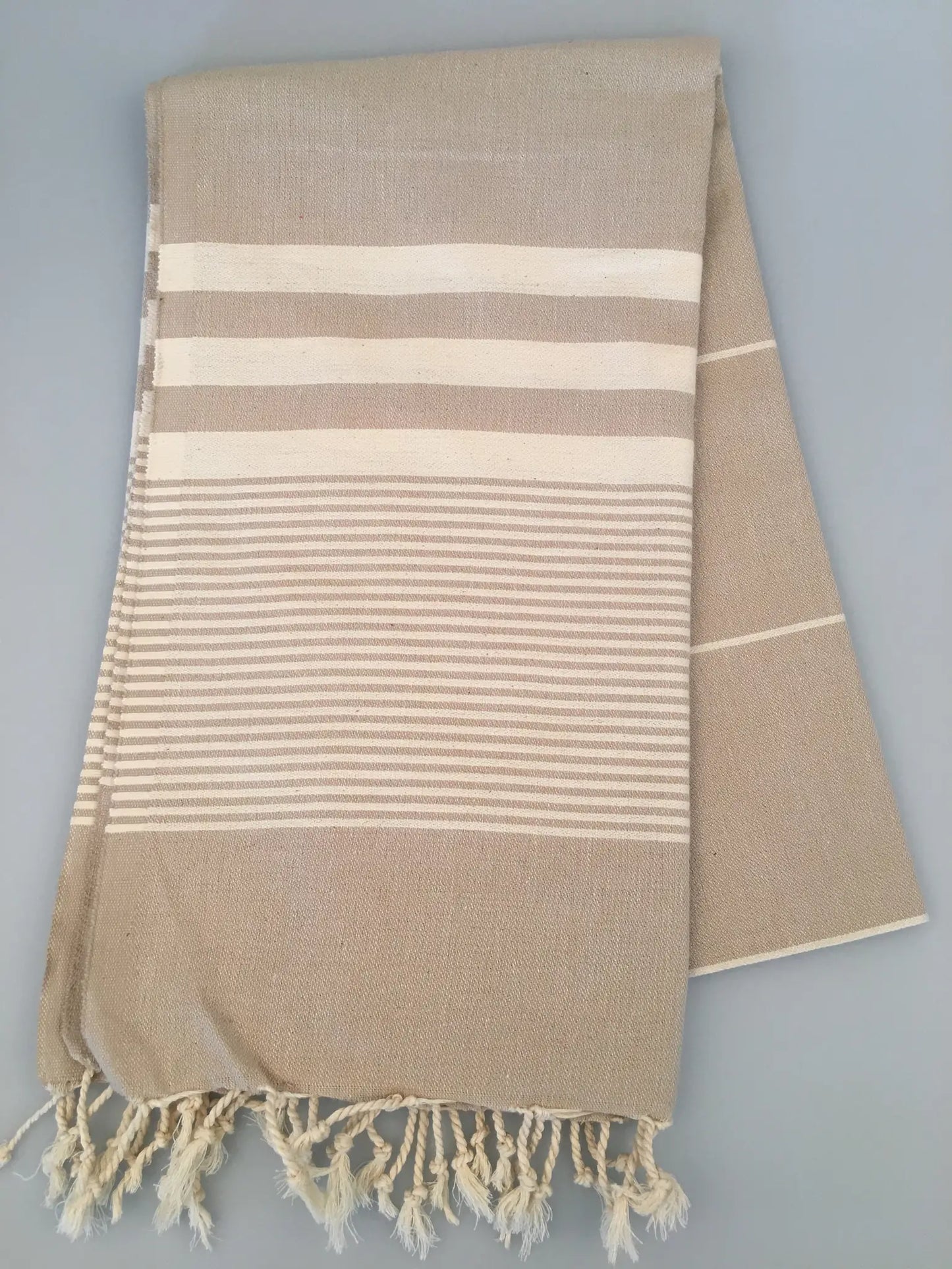Beige Turkish towel with white stripes and fringed ends, folded neatly on a light gray background.