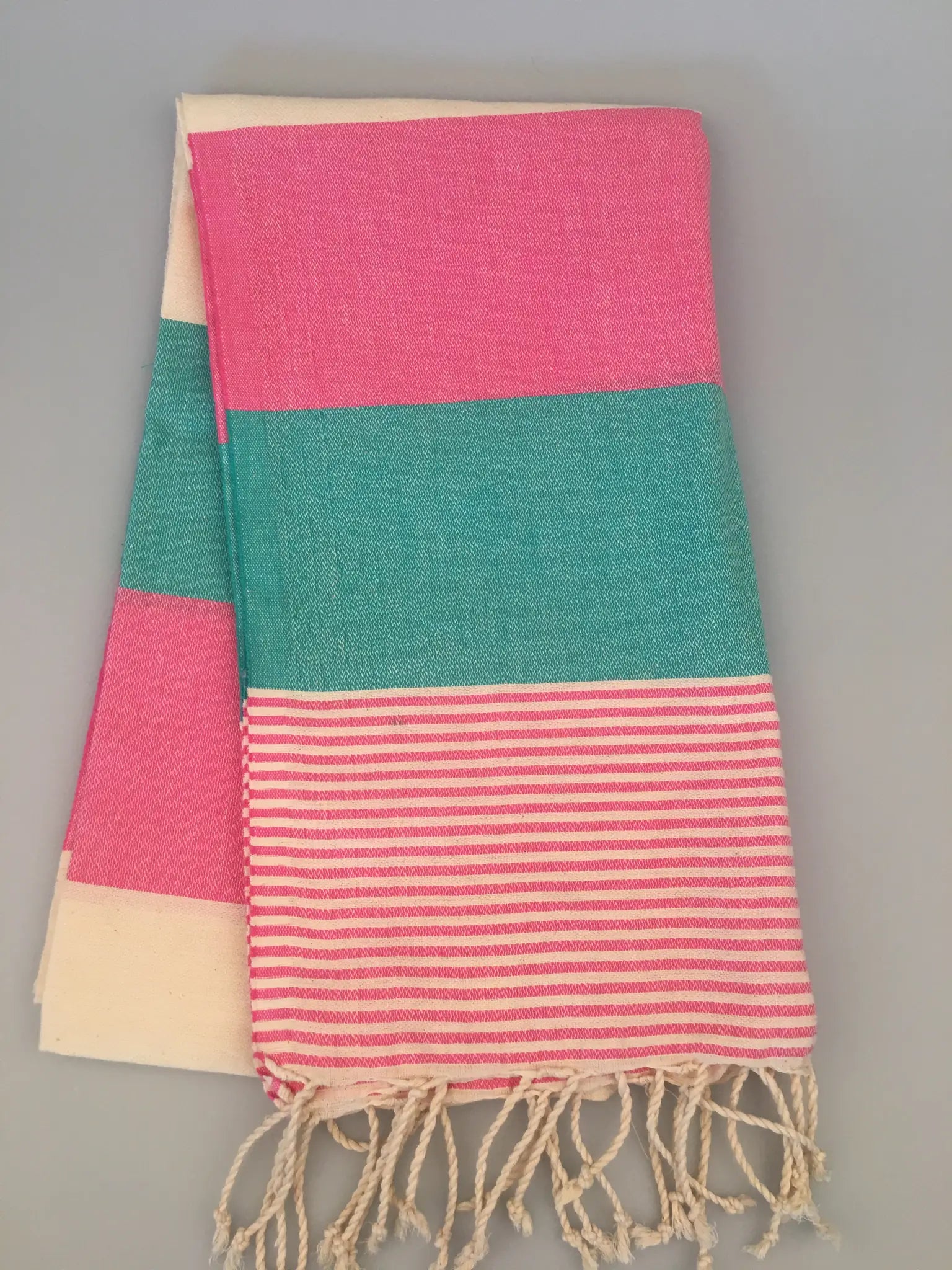 "Turquoise and pink striped Lagina Turkish towel with cream accents and fringed edges, folded on a gray background."