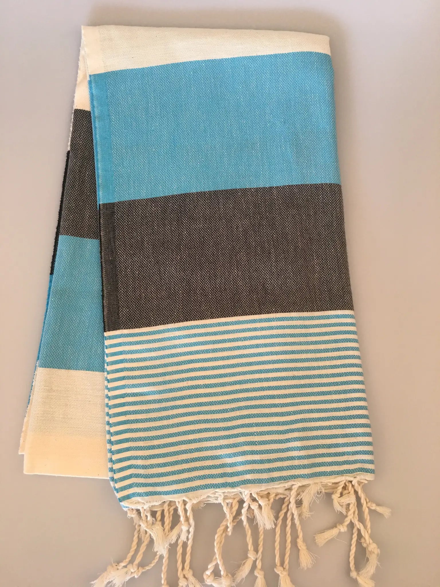 "Turquoise and gray striped Turkish towel with fringed edges, folded neatly on a light background."