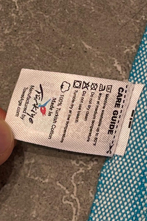 "White fabric label with black text, care instructions, and colorful logo, held against a textured blue background."