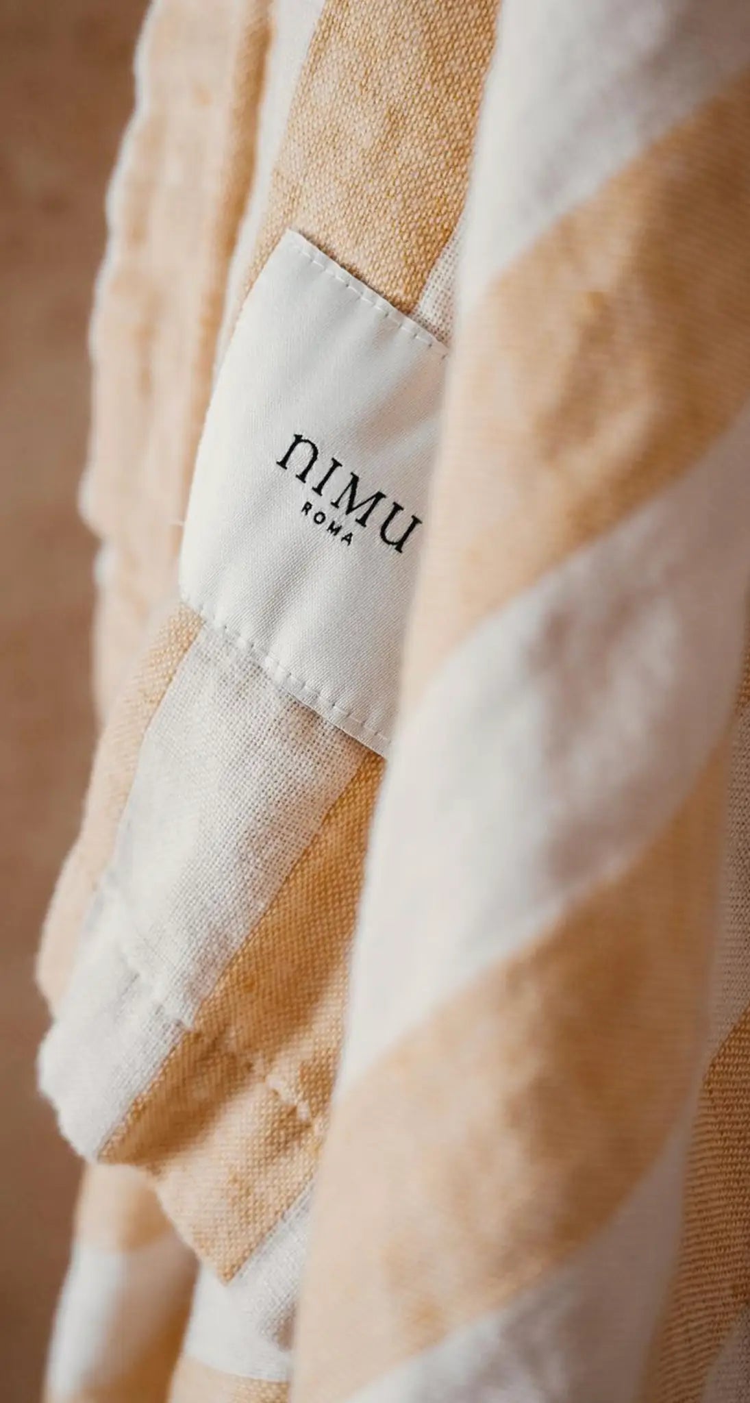 "Close-up of a cotton label with 'nimu' printed in black on a cream background, attached to a striped fabric in soft beige an