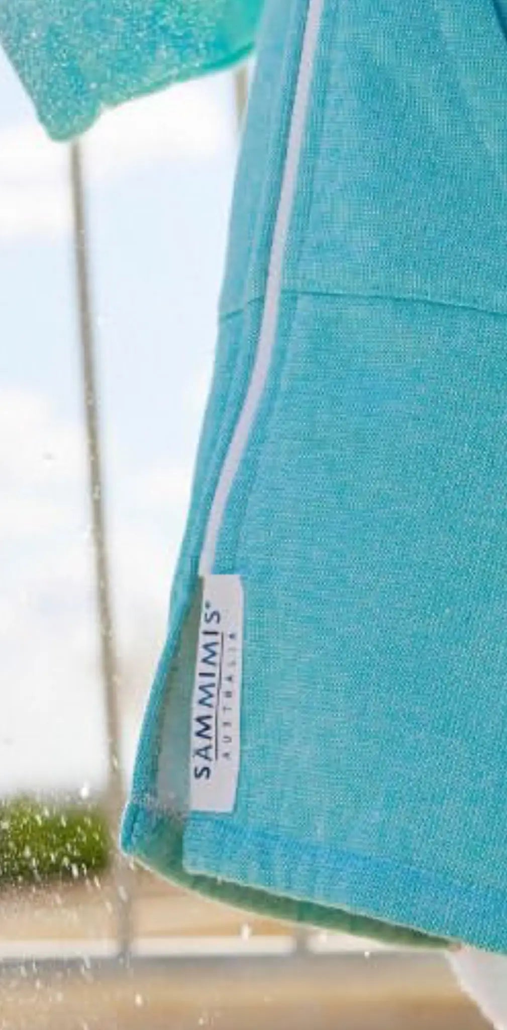 Light blue cotton label with white text reading "SAMMIMIS," attached to a turquoise garment, side view.