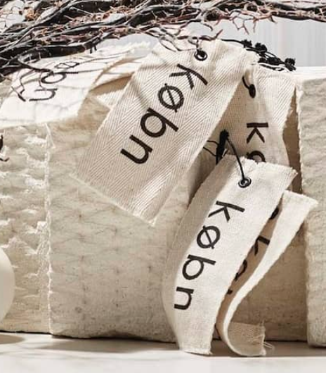 "Two cotton labels in cream color with black text, attached to a textured fabric surface, showcasing custom design."