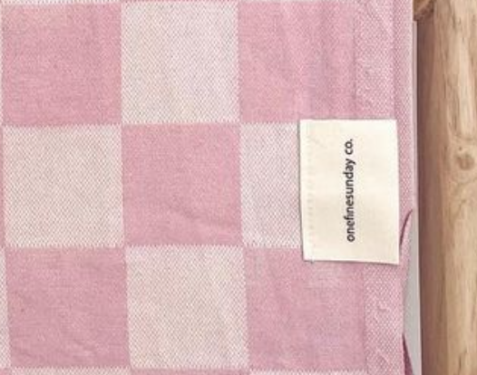 Pink and white checkered cotton fabric with a sewn label "onefinesunday co." at the edge, top-down view.