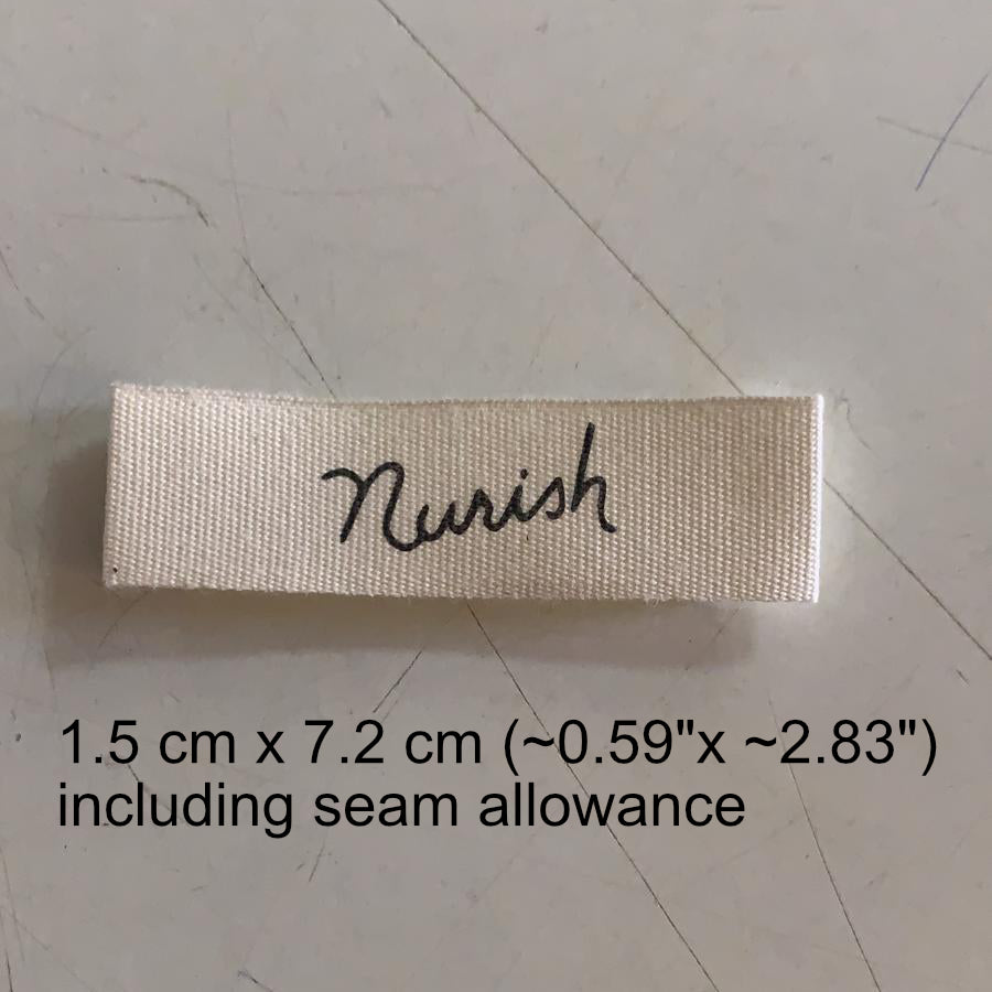 "Rectangular cotton label in cream with the word 'Nourish' in cursive black font, dimensions printed below."