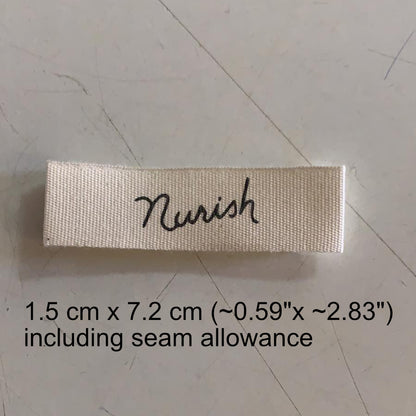 "Rectangular cotton label in cream with the word 'Nourish' in cursive black font, dimensions printed below."