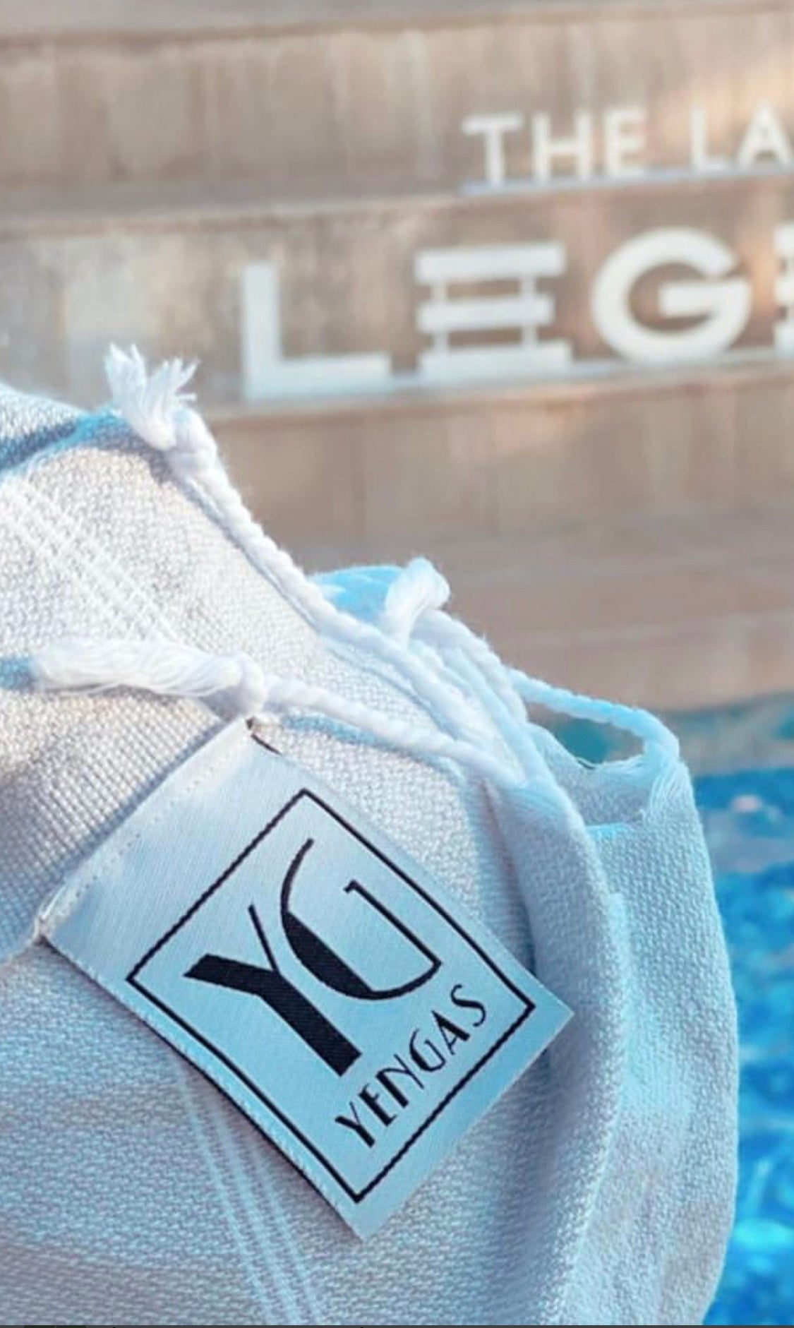 White cotton label with black text reading "YG YENGAS," partially visible against a blurred blue background.